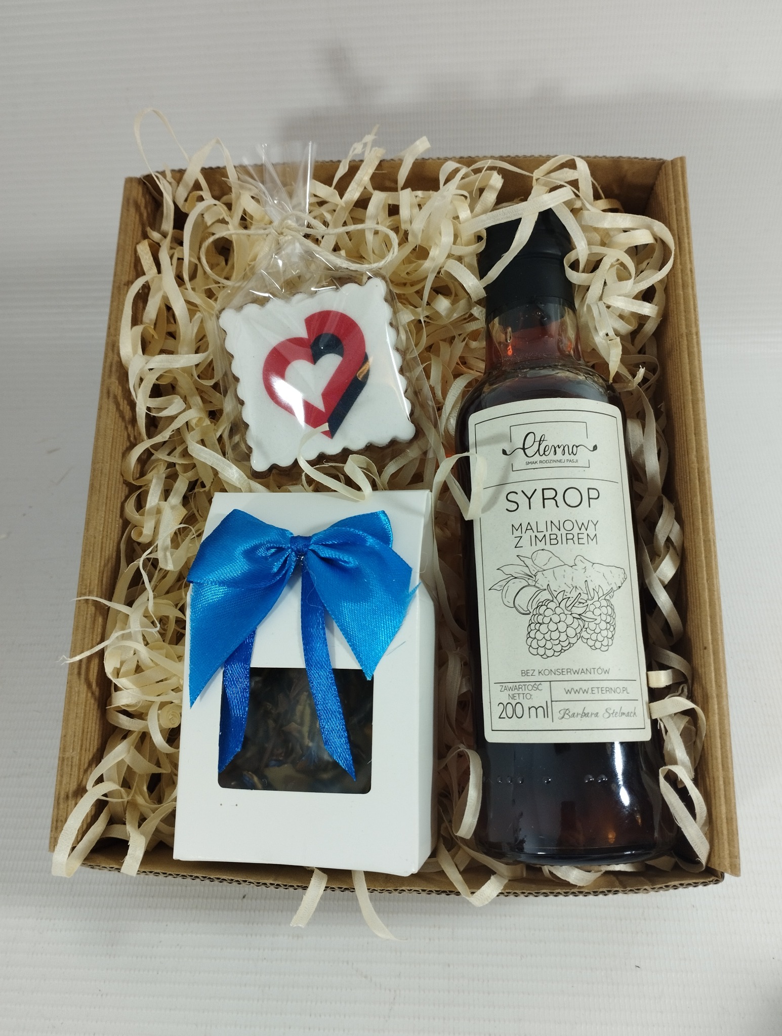 Kartonowy kosz prezentowy wypełniony ozdobnym siankiem, zawierający syrop malinowo-imbirowy, herbatę w białym pudełku z niebieską kokardą i piernik w kształcie serca.