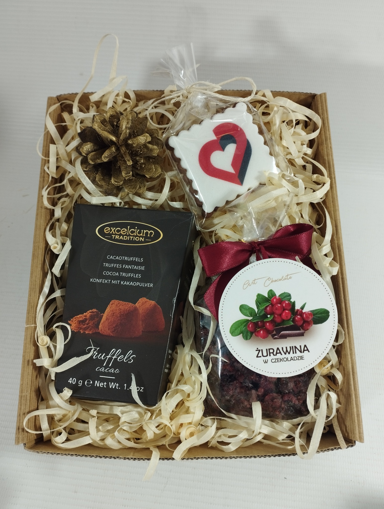 Elegancki kosz prezentowy z truflami, żurawiną w czekoladzie i piernikiem w kształcie serca, wypełniony dekoracyjnym siankiem w kartonowym pudełku.