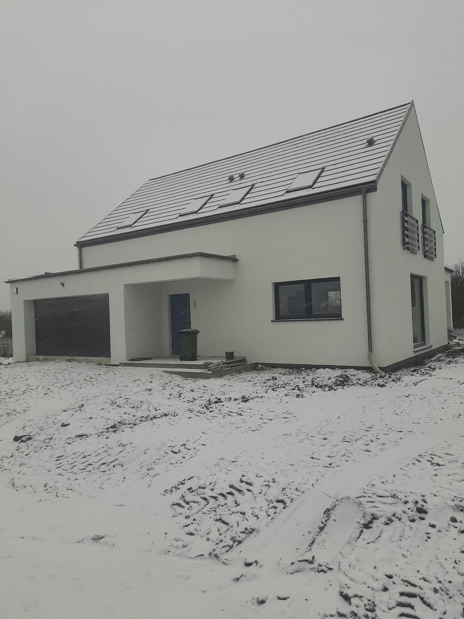 Nowoczesny, dwukondygnacyjny dom murowany z garażem w zimowej scenerii, pokryty śniegiem dach z oknami połaciowymi, biała elewacja, teren wokół domu zasypany śniegiem.