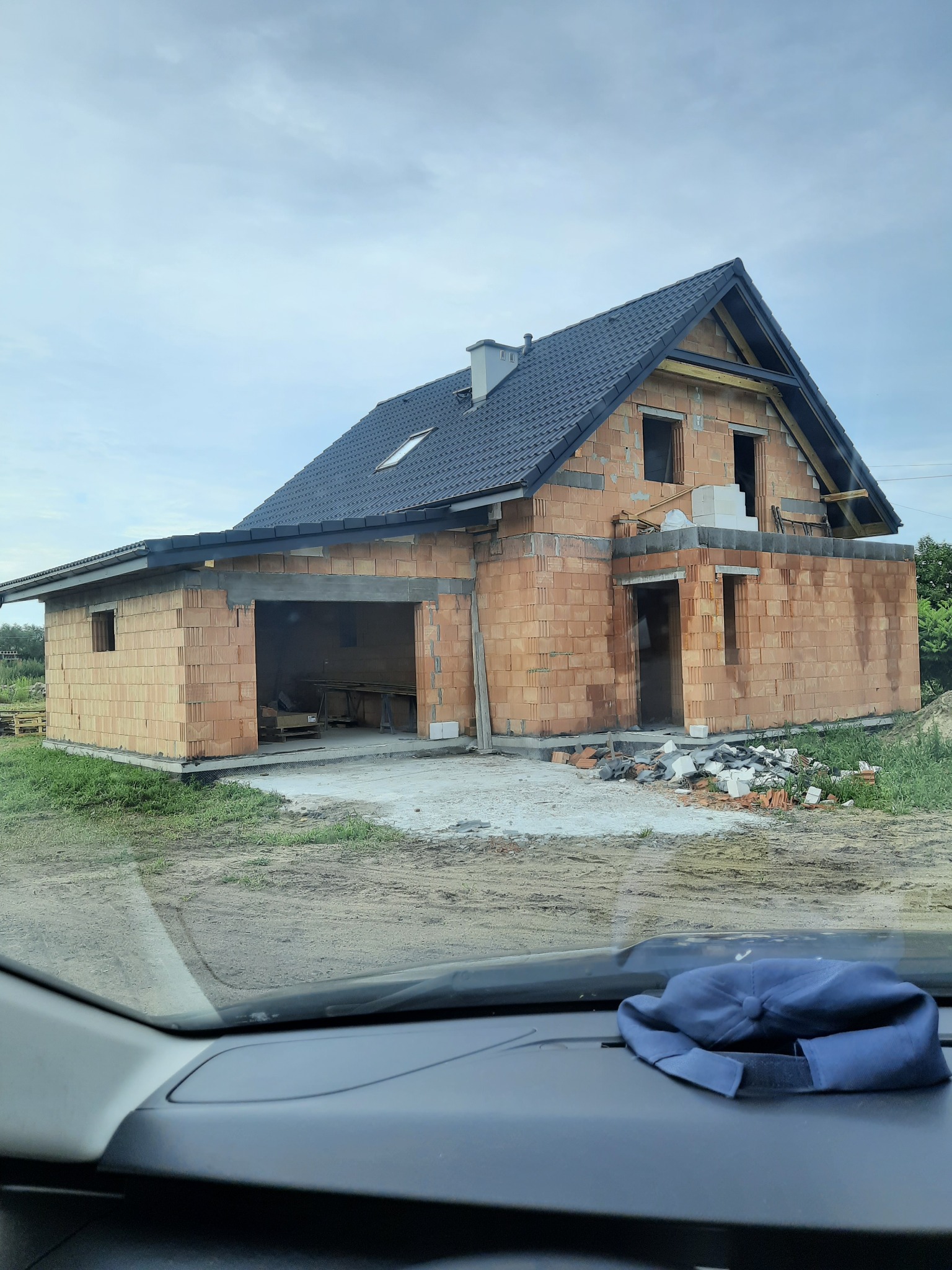 Surowy stan budowy domu z czerwonej cegły z dachem z ciemnej dachówki, widok z wnętrza samochodu z czapką na desce rozdzielczej.