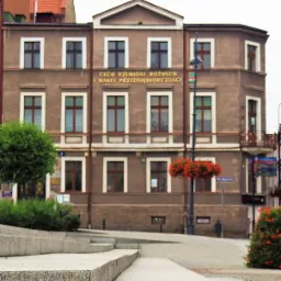 Dwupiętrowy budynek z napisem 'CECH RZEMIOSŁ RÓŻNYCH I MAŁEJ PRZEDSIĘBIORCZOŚCI', widoczny od dołu z perspektywy schodów, z elementami zieleni w tle.