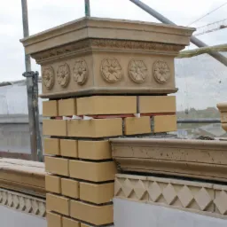 Wznoszenie muru z cegły klinkierowej z dekoracyjnym zwieńczeniem i podsypką. Widoczne rusztowania i żuraw budowlany w tle. Prace murarskie w toku.