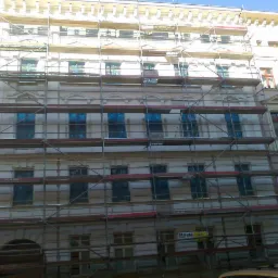 Elewacja kamienicy w trakcie renowacji, zabezpieczone okna, rusztowania na całej wysokości budynku, widoczny fragment chodnika i zaparkowanego auta.