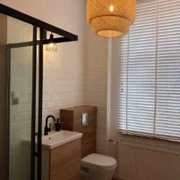 Nowoczesna łazienka z kabiną prysznicową typu walk-in, umywalką nablatową z czarną armaturą, podwieszaną toaletą, drewnianymi akcentami i dekoracyjną lampą wiszącą z plecionki.