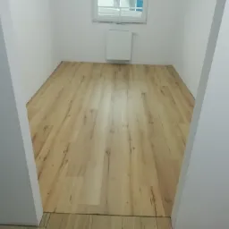 Nowo ułożone panele podłogowe w jasnym pomieszczeniu w Ostaszewie, widoczne gładkie, drewnopodobne panele i białe ściany, gotowe do dalszego wykończenia.