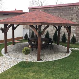 Wood M&A - Drewniana altana z czerwonym gontem bitumicznym, okrągłym stołem i krzesłami na kamiennym placu, otoczona trawnikiem i tujami.