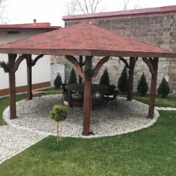 Drewniana altana z czerwonym gontem bitumicznym, okrągłym stołem i krzesłami na kamiennym placu, otoczona trawnikiem i tujami.