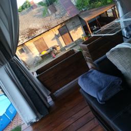 Wood M&A - Zadaszony taras drewniany w Opatowie z widokiem na podwórze, z meblami ogrodowymi i zasłonami, w tle zabudowania gospodarcze.