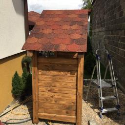 Wood M&A - Drewniana obudowa studni z czerwoną, łuskową dachówką, żółtym wężem ogrodowym u podstawy i aluminiową drabiną w tle, na kamiennym podłożu.