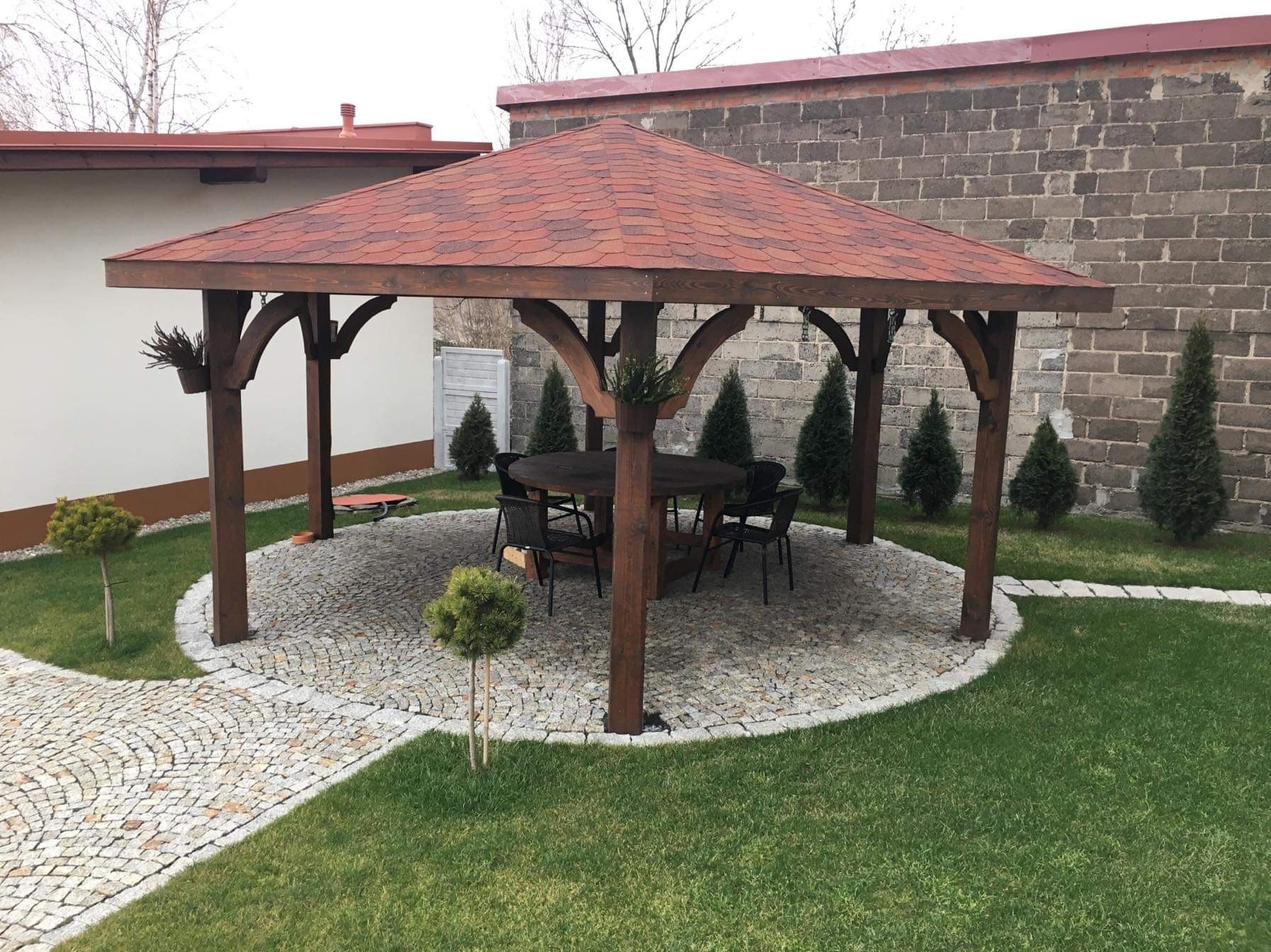 Drewniana altana z czerwonym gontem bitumicznym, okrągłym stołem i krzesłami na kamiennym placu, otoczona trawnikiem i tujami.