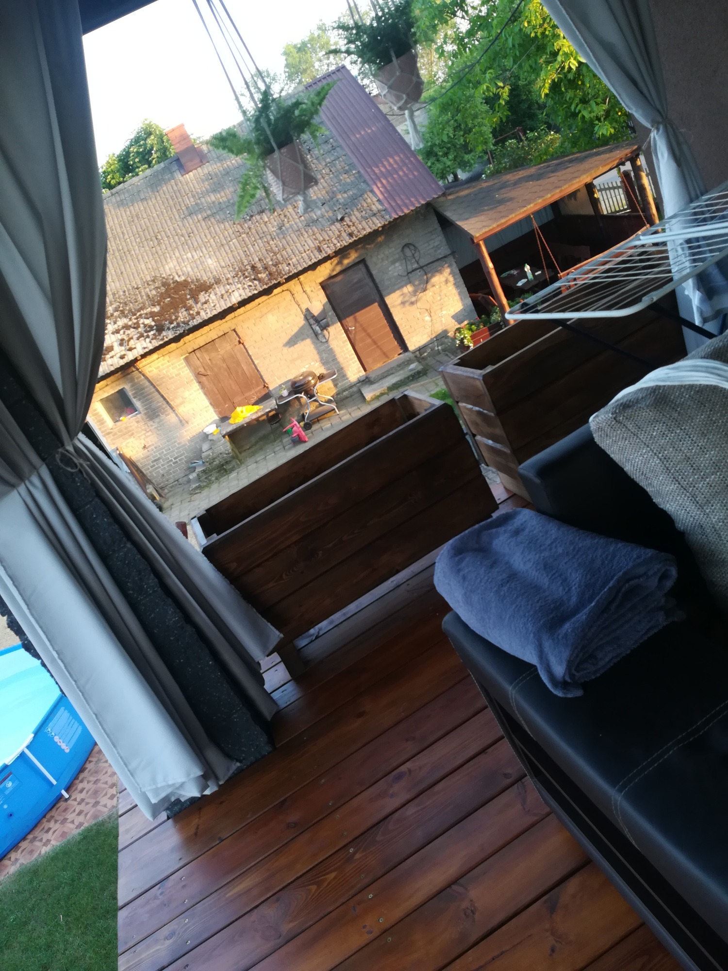 Zadaszony taras drewniany w Opatowie z widokiem na podwórze, z meblami ogrodowymi i zasłonami, w tle zabudowania gospodarcze.