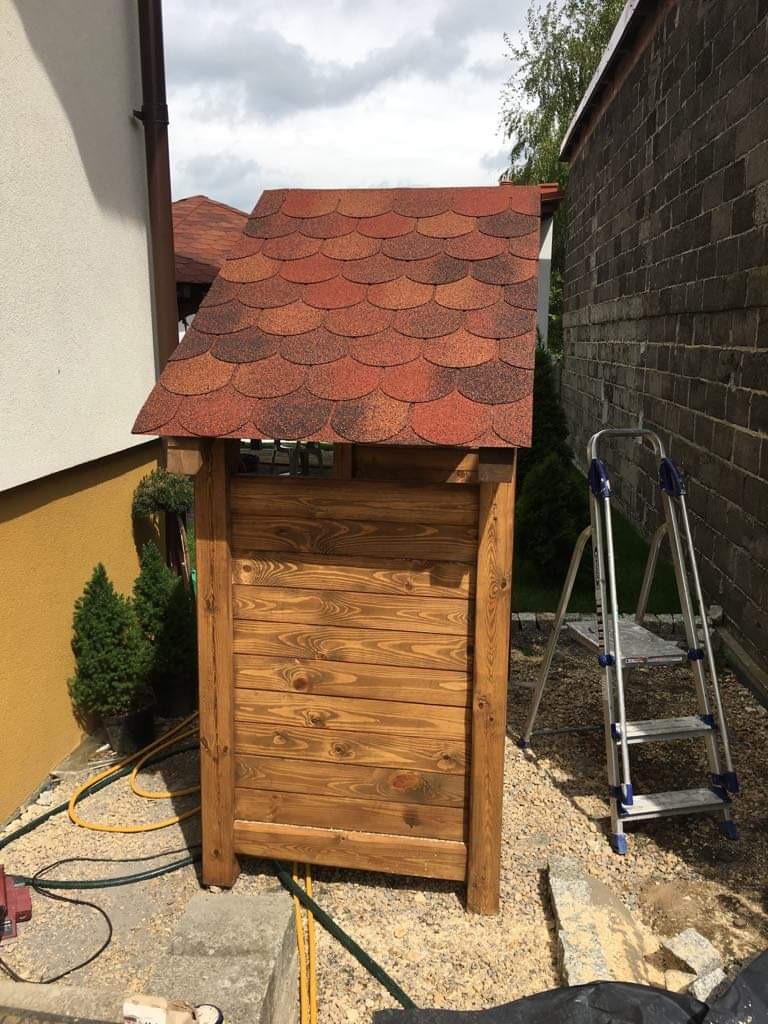 Drewniana obudowa studni z czerwoną, łuskową dachówką, żółtym wężem ogrodowym u podstawy i aluminiową drabiną w tle, na kamiennym podłożu.