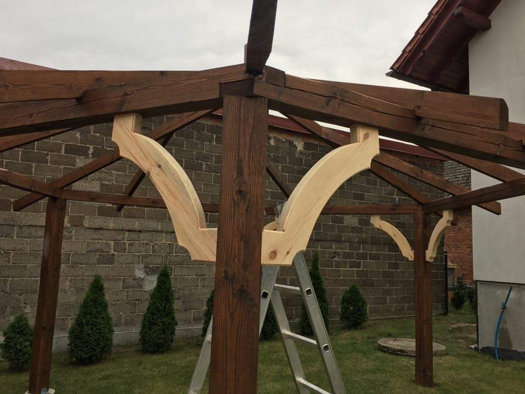 Konstrukcja z ciemnego drewna w trakcie montażu, widoczne jasne, surowe łuki podtrzymujące, drabina oparta o słup, tło z cegły i zieleni.