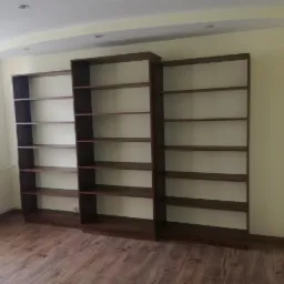 Trzy brązowe regały na książki bez zawartości, ustawione w rzędzie na drewnianej podłodze w jasnym pokoju z białym sufitem i jasnymi ścianami.