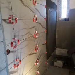 Ściana w trakcie układania płytek imitujących marmur, z użyciem czerwonych klipsów i czarnego sznurka do poziomowania, widoczne okno i surowa ściana w tle.