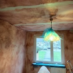 Łukasz Markowski - Pokój w trakcie malowania ścian na brązowo, zabezpieczone okno i grzejnik folią, widoczny sufit z niedomalowanymi pasami, nietypowa lampa z abażurem w kształcie kwiatu.