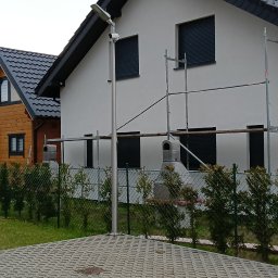 Łukasz Markowski - Dwupiętrowy dom z szarą elewacją, czarnym dachem i zewnętrznymi roletami w oknach, częściowo zasłonięty rusztowaniem, widoczny zza metalowego ogrodzenia i chodnika z betonowej kostki. Obok widoczny...