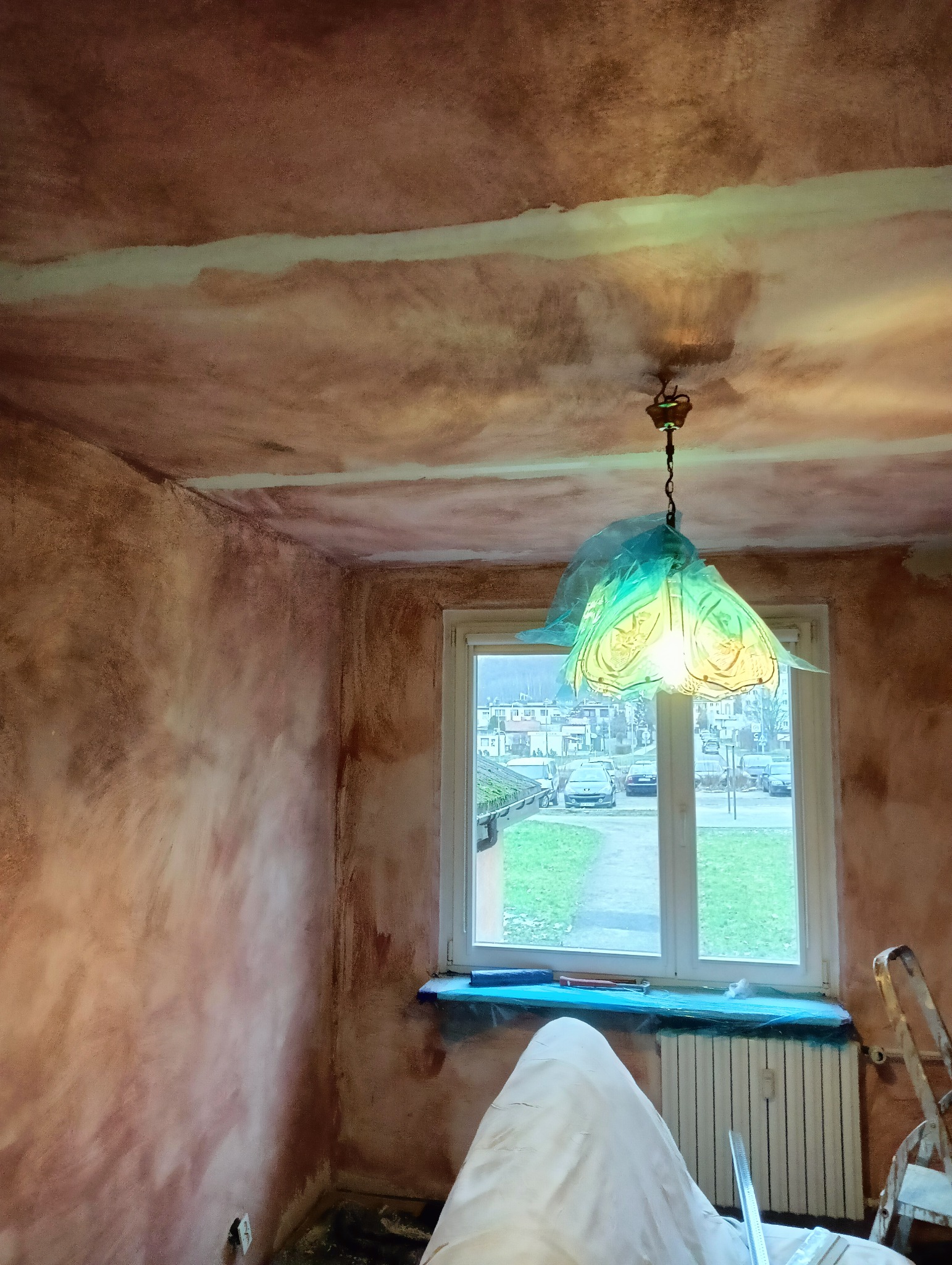 Pokój w trakcie malowania ścian na brązowo, zabezpieczone okno i grzejnik folią, widoczny sufit z niedomalowanymi pasami, nietypowa lampa z abażurem w kształcie kwiatu.