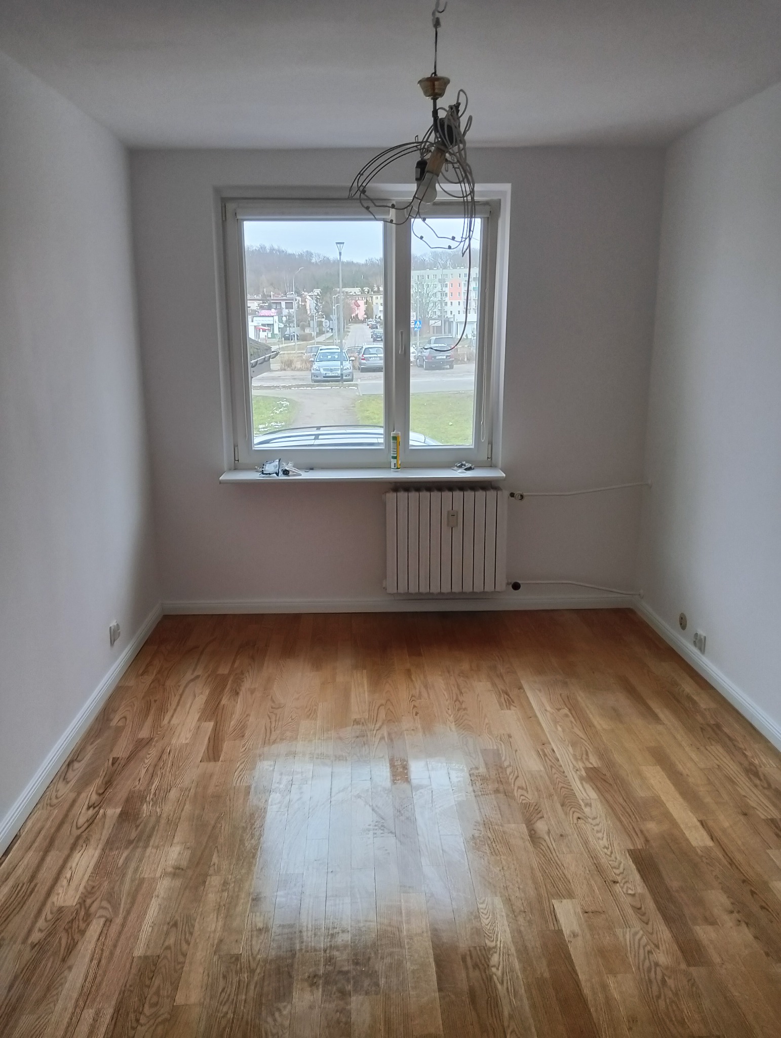 Pusty pokój z drewnianą podłogą, białymi ścianami i grzejnikiem pod oknem, widoczne kable od lampy wiszącej.