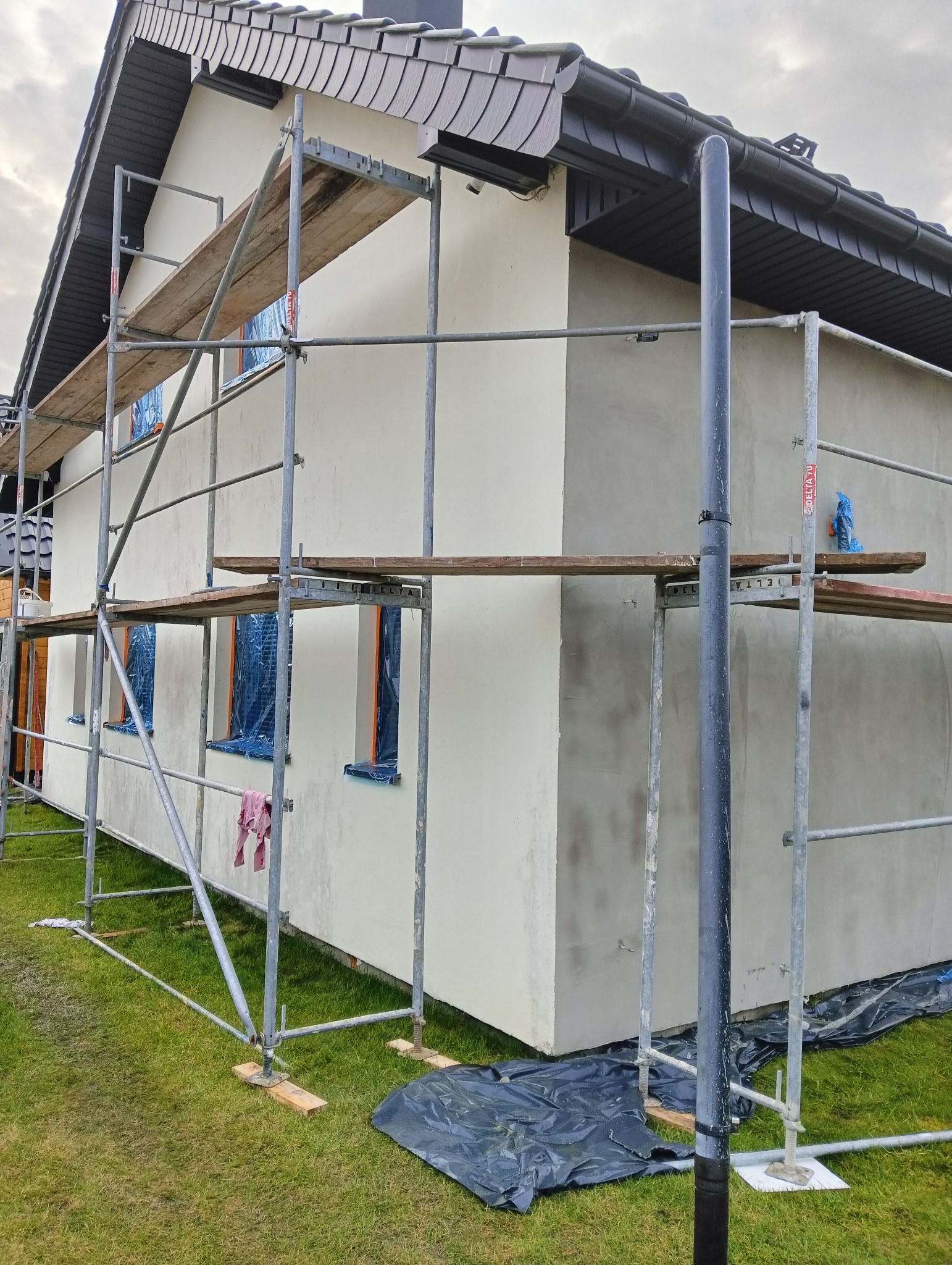 Elewacja domu w trakcie prac wykończeniowych, z widocznymi rusztowaniami, zabezpieczonymi oknami i fragmentami świeżo nałożonego tynku.