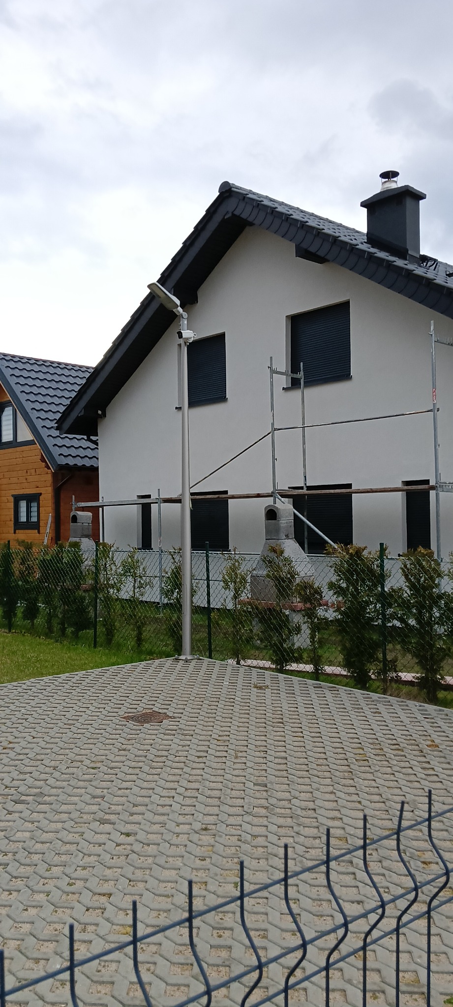 Dwupiętrowy dom z szarą elewacją, czarnym dachem i zewnętrznymi roletami w oknach, częściowo zasłonięty rusztowaniem, widoczny zza metalowego ogrodzenia i chodnika z betonowej kostki. Obok widoczny...