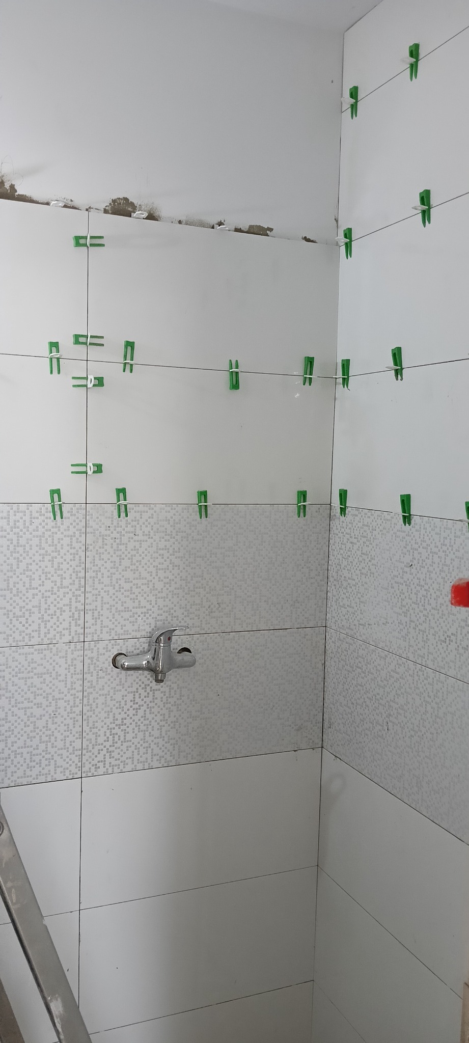 Białe płytki z mozaikowym pasem w trakcie układania w rogu łazienki, widoczne zielone klipsy poziomujące i sznurki, na ścianie bateria umywalkowa, ślady zaprawy na suficie.