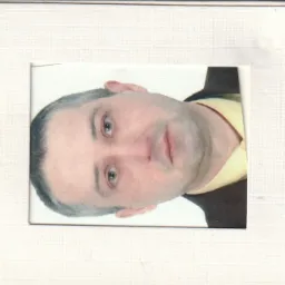 Zdjęcie portretowe mężczyzny w brązowej marynarce i żółtej koszuli, umieszczone w białej ramce o fakturze płótna.