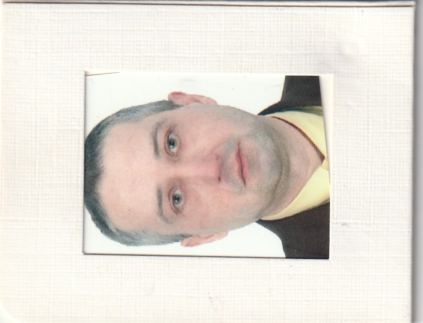 Zdjęcie portretowe mężczyzny w brązowej marynarce i żółtej koszuli, umieszczone w białej ramce o fakturze płótna.