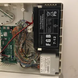 Wnętrze otwartej obudowy centrali alarmowej z widoczną płytą główną, akumulatorem FGB 7-12 oraz transformatorem NORATEL LF688-1818, zasilanie 230V.