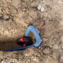 Instalacjekompleksowo.pl - Event Plus - Niebieski karbowany przewód i czarny kabel elektryczny zabezpieczony czerwoną taśmą, ułożone w wykopie ziemnym.