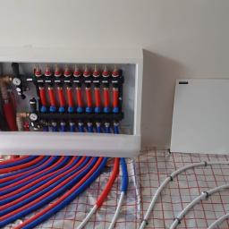 Instalacjekompleksowo.pl - Event Plus - Rozdzielacz ogrzewania podłogowego w białej szafce z czerwono-niebieskimi rurami na folii grzewczej, widoczny fragment instalacji elektrycznej i betonowego słupa.