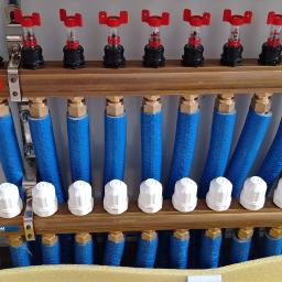 Instalacjekompleksowo.pl - Event Plus - Rozdzielacz ogrzewania podłogowego Ferro z niebieskimi rurami w szafce, widoczne zawory z czerwonymi pokrętłami i białe głowice termostatyczne.