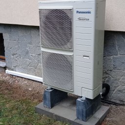 Instalacjekompleksowo.pl - Event Plus - Zewnętrzna jednostka klimatyzacji Panasonic Inverter, zamontowana na betonowych blokach przy ścianie budynku z elewacją z kamienia łupanego.