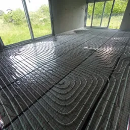 Rozłożone rury ogrzewania podłogowego na folii izolacyjnej w surowym wnętrzu z dużymi oknami wychodzącymi na zieleń.