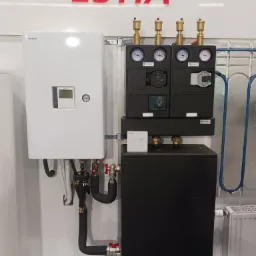 Kompletna instalacja grzewcza z pompą ciepła Toshiba Estia, widoczne elementy sterujące, manometry Afriso, rury izolowane i grzejnik panelowy.