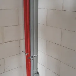 Dwie czerwone rury i szara rura kanalizacyjna przymocowane do ściany z bloczków betonowych, widoczne obejmy mocujące i fragment posadzki.
