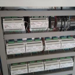 Instalacjekompleksowo.pl - Event Plus - Rozdzielnica elektryczna z modułami sterującymi, widoczne przewody i złączki, wewnątrz szarej szafy sterowniczej.