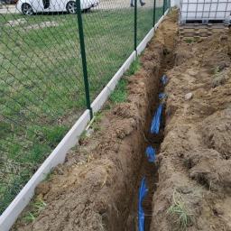 Instalacjekompleksowo.pl - Event Plus - Wkopane w ziemi rury w niebieskiej osłonie, przygotowane do podłączenia do zbiornika na wodę, widoczne przy ogrodzeniu z siatki.