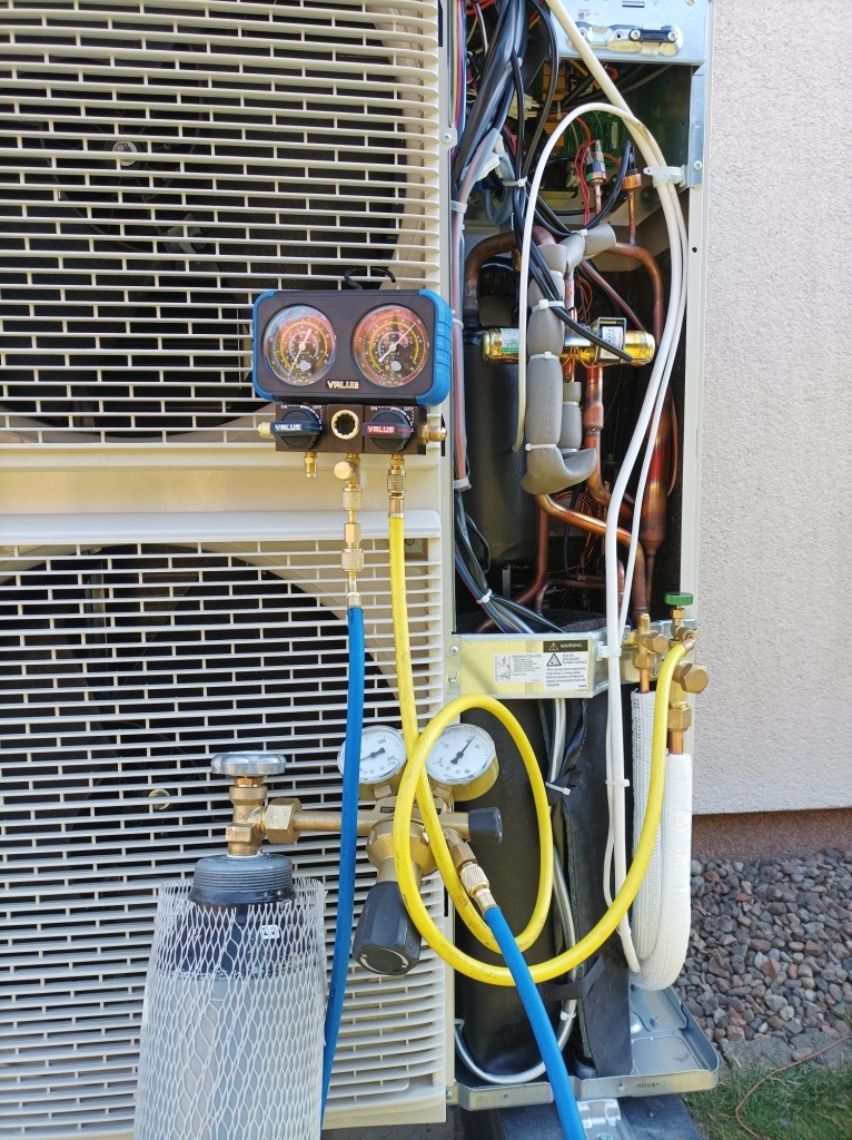 Podłączenie manometrów do pompy ciepła podczas serwisu, widoczna butla z czynnikiem chłodniczym i przewody w kolorach niebieskim i żółtym.