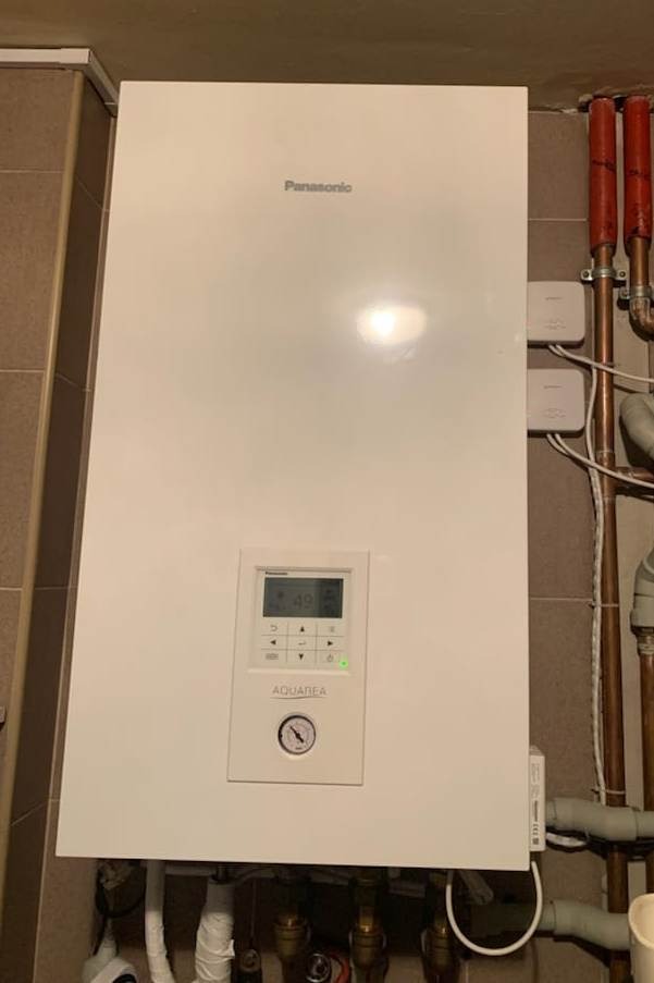 Biała pompa ciepła Panasonic Aquarea na ścianie z beżowymi płytkami, widoczne rury miedziane i plastikowe podłączenia hydrauliczne.