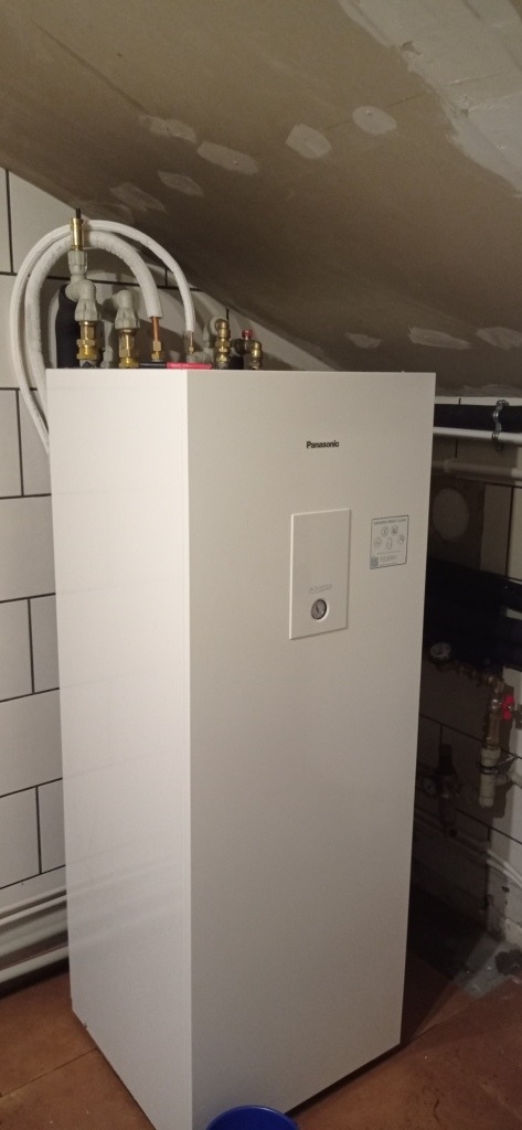 Biała jednostka wewnętrzna pompy ciepła Panasonic, z widocznymi przyłączami hydraulicznymi u góry, zamontowana w pomieszczeniu z białymi płytkami i surowym sufitem.