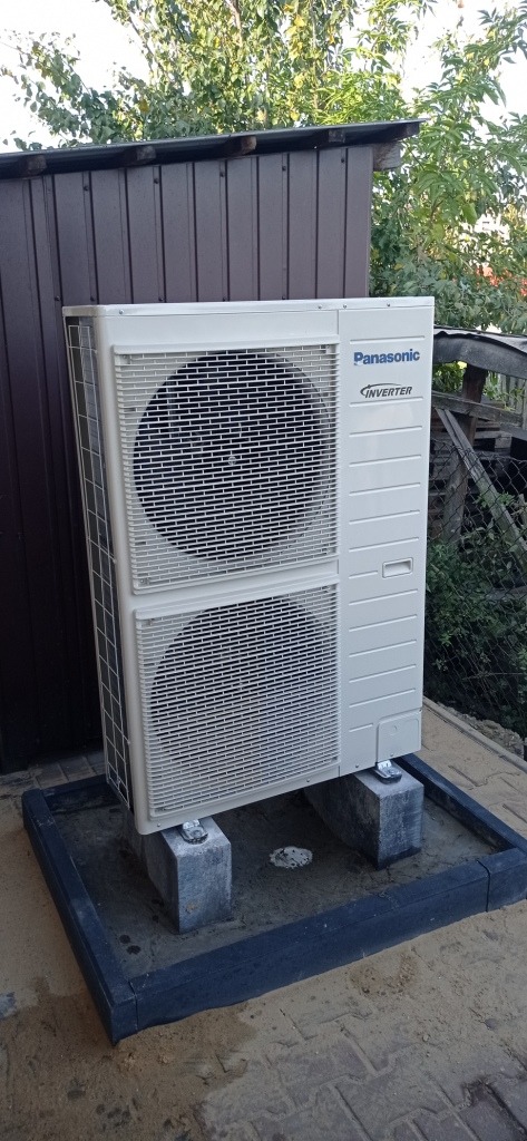 Zewnętrzna jednostka klimatyzacji Panasonic Inverter, ustawiona na betonowych bloczkach w czarnej obudowie, na tle brązowej ściany budynku i zieleni.