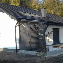 Nowoczesny, biały dom jednorodzinny z ciemną dachówką i elementami drewnianymi, widok z zewnątrz, z klimatyzatorem na ścianie i elementami w trakcie wykańczania wokół domu.