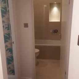 Widok na wyremontowaną łazienkę z toaletą i wanną zza otwartych drzwi, z widoczną ścianą w tropikalne wzory w sąsiednim pomieszczeniu.