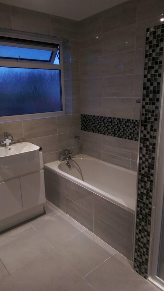Wykończona łazienka z beżowymi płytkami, białą wanną i umywalką, oraz dekoracyjnym pasem mozaiki nad wanną i przy kabinie prysznicowej; za oknem widoczny deszcz.