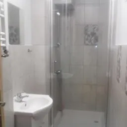 Niewielka łazienka z kabiną prysznicową z transparentnego szkła, umywalką narożną i grzejnikiem drabinkowym, wykończona jasnymi płytkami z dekoracyjnymi wstawkami.