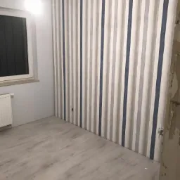 Pomalowane ściany w biało-szaro-niebieskie pasy w pokoju z jasną podłogą panelową, okno z ciemnymi zasłonami po lewej stronie, widoczne gniazdko elektryczne na ścianie.