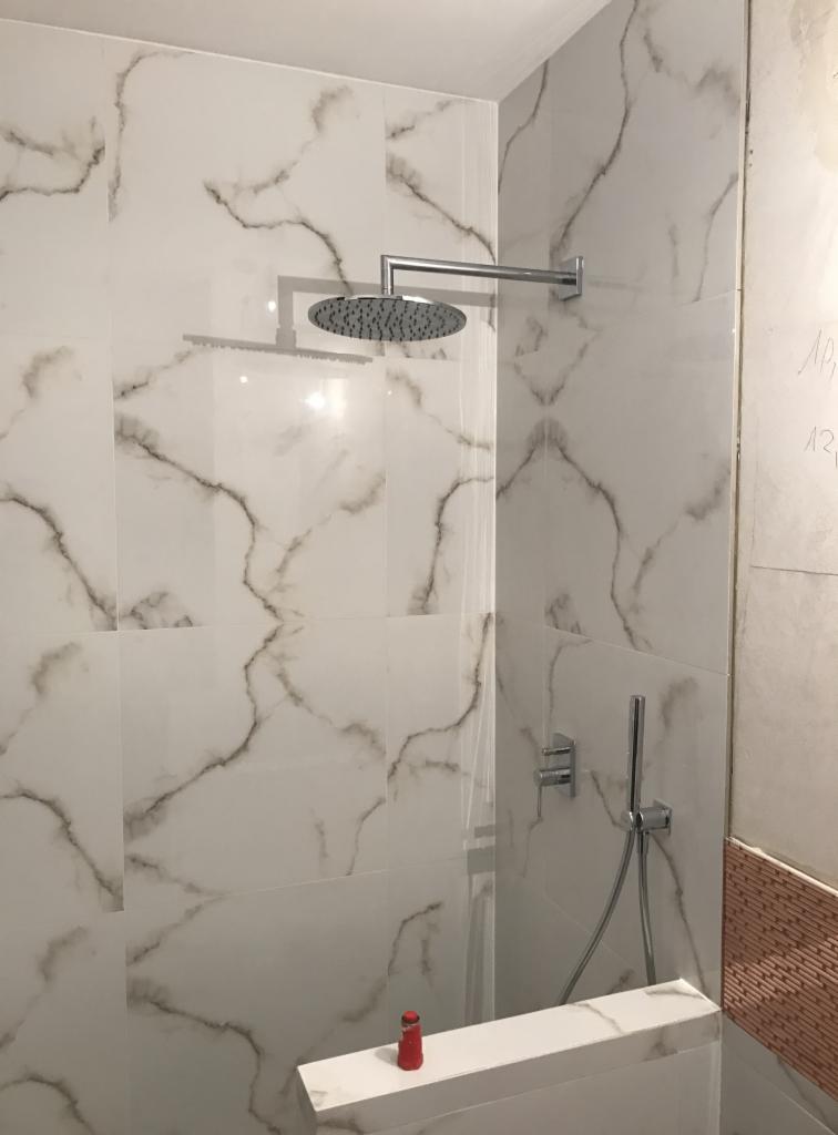 Kabina prysznicowa z deszczownicą, wykończona białymi płytkami imitującymi marmur z szarymi żyłami, widoczna czerwona buteleczka na obudowie wanny.