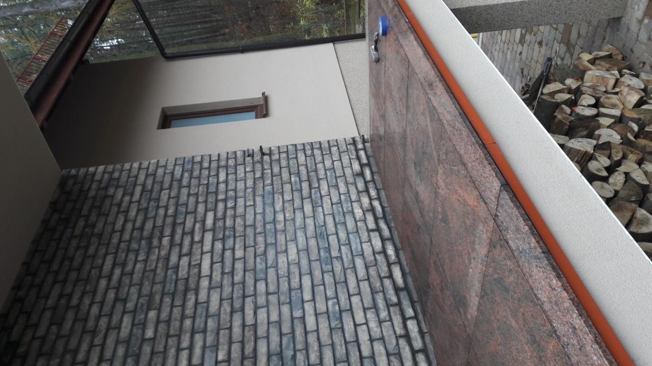 Wykończona elewacja zewnętrzna z imitacji cegły, widoczne okno, fragment dachu oraz składowane drewno.