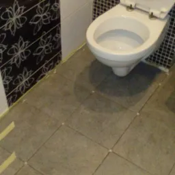 Toaleta z otwartą klapą, częściowo wykończona glazurą na ścianach i szarymi płytkami podłogowymi z krzyżykami dystansowymi, widoczne taśmy malarskie przy ścianie.